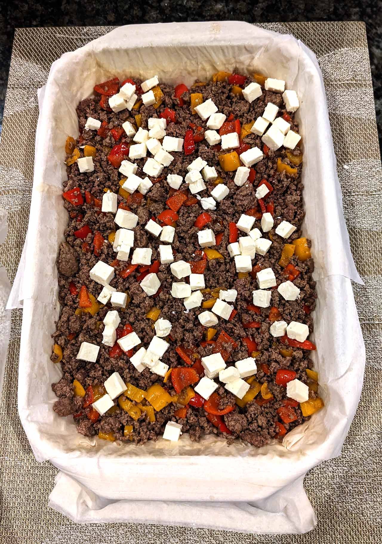 Baking dish lined with raw phyllo dough and topped with sautéed ground beef, peppers, and feta cheese cubes