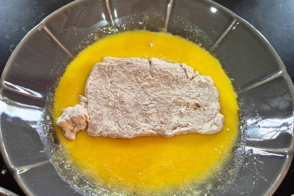 Pork cutlet coated in flour and laying in a bowl of raw scrambled eggs
