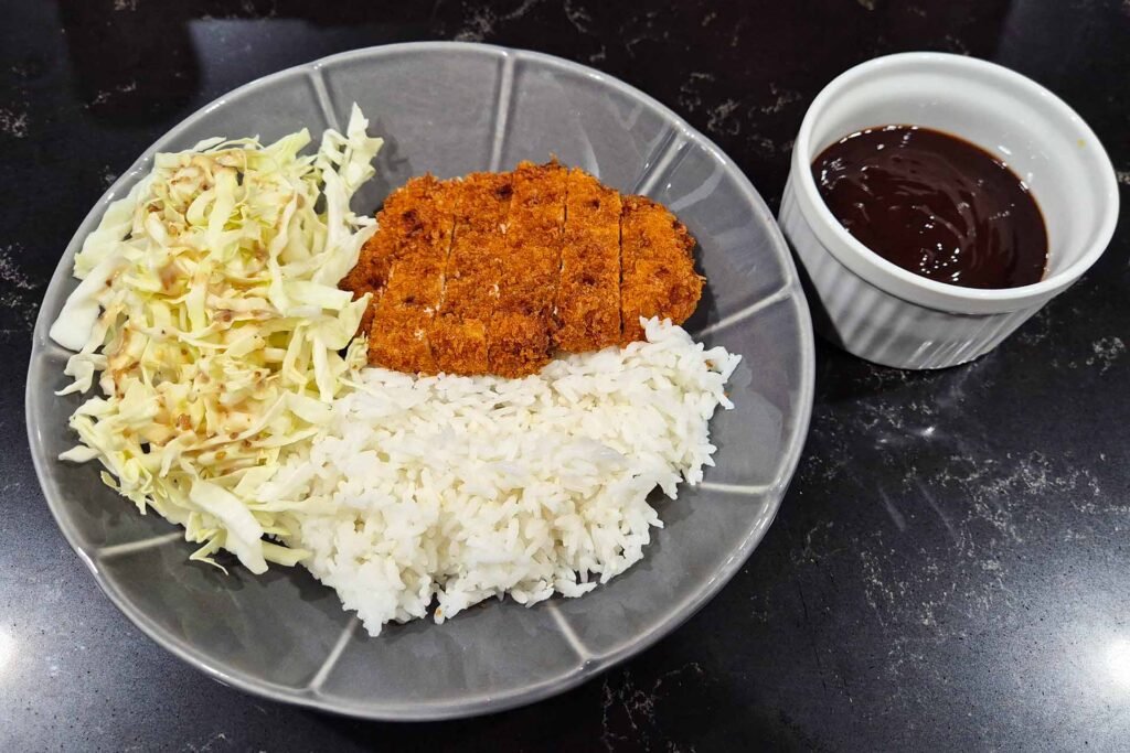 A plate of pork tonkatsu, cabbage salad, and white rice with sauce on the side